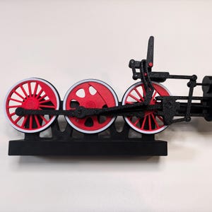May include: A black and red mechanical train wheel assembly featuring three circular wheels with red spokes and white rims, connected by a black linkage system mounted on a rectangular black base, showcasing intricate moving parts and gears.
