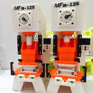 Puede incluir: Dos prensas mecánicas blancas y naranjas con el texto MF26-125 impreso en la parte superior. Cada máquina cuenta con un dial circular tipo reloj con una sola manecilla, un mecanismo de presión naranja central y un pequeño panel de control.