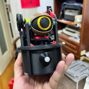 Puede incluir: Un dispositivo mecánico con una esfera central marrón con un iris circular amarillo, montado sobre una base cilíndrica negra. La estructura incluye engranajes negros, rojos y verdes expuestos, un pequeño sensor blanco y piezas de plástico.