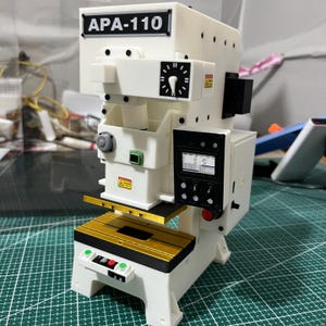 May include: A white scale model of an industrial power press machine labeled APA-110. The unit features a gold-colored work bed, a black control panel with a digital screen, a circular gauge, and various small red, green, and gray buttons and switches.