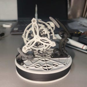 May include: A mechanical clock movement model featuring a white circular base with a black ribbed side. A white three-spoke gear assembly sits atop the base, supported by a black curved structural arm and smaller interlocking grey gears.