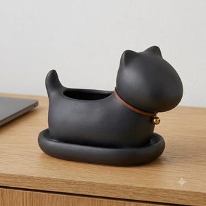 May include: A matte black ceramic planter shaped like a stylised dog with a smooth finish. The planter features a brown leather-like collar around the neck with a small, round, metallic gold-toned bell attached to the front of the collar.
