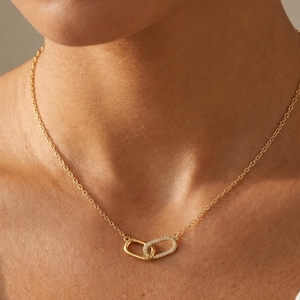 May include: A gold-toned chain necklace featuring two interlocking oval-shaped links as a central pendant. One of the oval links is plain gold-toned metal, while the other is encrusted with small, clear, sparkling stones along its entire surface.
