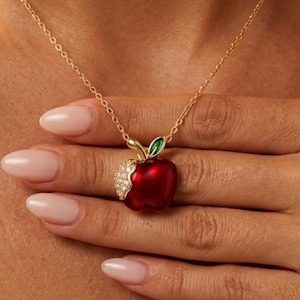 Op de afbeelding: Een gouden ketting met een hanger in de vorm van een rode appel. De appel heeft een klein groen blaadje aan de bovenkant en een gedeelte dat is ingelegd met heldere, fonkelende steentjes aan één kant. De hanger hangt aan een fijne goudkleurige ketting.
