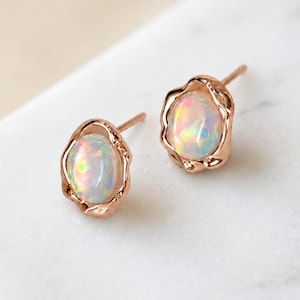 May include: A pair of stud earrings featuring oval-shaped opals with iridescent flashes of colour. Each opal is set within a rose gold metal frame that has an irregular, organic, and textured edge. The earrings include thin posts for attachment.