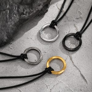 Puede incluir: Cuatro colgantes circulares en collares de cordón negro. Los colgantes incluyen un anillo plateado pulido, un anillo negro mate, un anillo plateado desgastado con marcas grabadas y un anillo dorado con un acabado liso y reflectante.