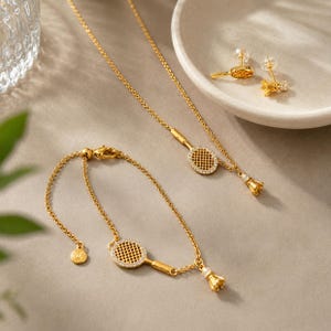 Könnte beinhalten: Ein goldfarbenes Schmuckset bestehend aus einer Halskette, einem Armband und einem Paar Ohrsteckern. Jedes Teil zeigt ein Tennisschläger-Motiv mit kleinen klaren Steinen und einem kleinen Federball-Anhänger an den Kettengliedern.