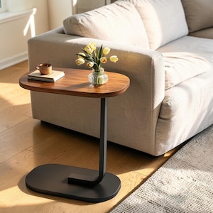 Può includere: Un tavolino a forma di C con piano ovale in legno scuro e base in metallo nero opaco. Sul tavolo sono presenti un vaso in vetro trasparente a coste con tulipani gialli, una piccola tazza in ceramica marrone e una pila di due libri.