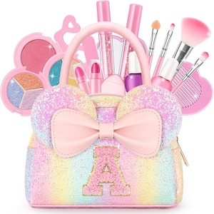 Peut inclure: Un sac à main pailleté aux tons pastel avec un grand nœud et deux oreilles arrondies sur le dessus, orné de la lettre A au centre. Le sac contient divers articles de maquillage, notamment des rouges à lèvres, des pinceaux et des palettes.