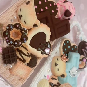 May include: Two phone cases decorated with miniature faux sweets including chocolate bars, heart-shaped cookies with pink polka dots, a donut with sprinkles, a bear-shaped cookie, a square biscuit with the word BISCUIT, and small pearls.