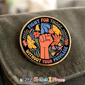 May include: A round enamel pin with a gold-toned metal edge featuring a central raised fist graphic surrounded by colorful flowers and leaves on a black background. The text "FIGHT FOR THOSE WITHOUT YOUR PRIVILEGE" is printed in a circular arc.