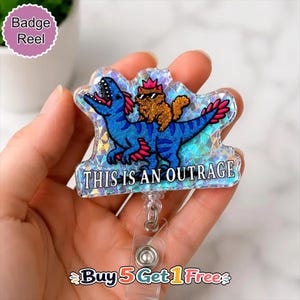 May include: A holographic badge reel featuring a blue dinosaur with pink accents and a brown cat wearing a pink crown and sunglasses on its back. The text "THIS IS AN OUTRAGE" is printed in white at the bottom of the acrylic piece.