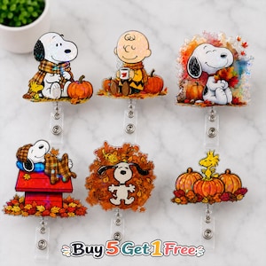 May include: Six acrylic badge reels featuring Peanuts characters in autumn themes. Designs include Snoopy with pumpkins, Charlie Brown with a mug, Snoopy in leaves, Snoopy on a doghouse, Snoopy in foliage, and Woodstock on pumpkins. Text reads Buy 5 Get 1 Free.