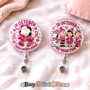 May include: Two circular badge reels with a holographic glitter finish. The left reel features Snoopy in pink boxing gear with a pink ribbon and the text IN OCTOBER WE WEAR PINK. The right reel shows Snoopy, Charlie Brown, Lucy, and Linus in pink winter clothing.