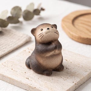 Könnte beinhalten: Eine kleine, sitzende Otter-Figur aus Keramik mit einer texturierten, braunen Farbverlauf-Oberfläche. Der Otter hat ein hellbraunes Gesicht, einen dunkelbraunen Körper und kleine Ohren, wobei die Vorderpfoten vor der Brust zusammengehalten sind.