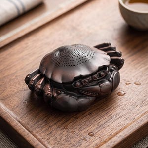 以下が含まれることがあります： 濃い茶色の粘土で作られたカニの形をした茶こし。甲羅の上部には茶葉を濾すための小さな円形の穴が詳細なパターンで施されており、本体にはリアルに成形された脚とハサミがマットな質感で表現されています。