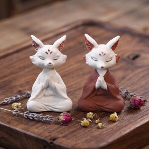Puede incluir: Dos figuras de zorro en posición de meditación sentada con las manos juntas. Un zorro es blanco y el otro es de color marrón rojizo. Ambos tienen los ojos cerrados, orejas puntiagudas con detalles internos rojizos y marcas en la frente.