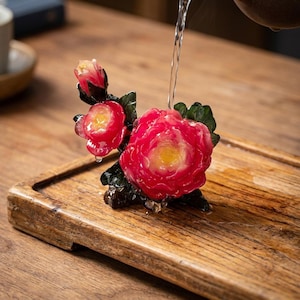 May include: A decorative tea pet sculpture shaped like a blooming red peony flower with a yellow centre and dark green leaves. Water is being poured over the glossy, translucent red petals, causing droplets to bead on the surface of the flower.