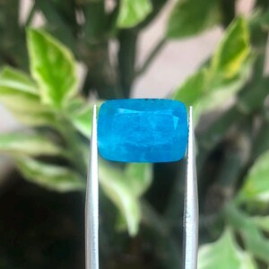 May include: A cushion-cut, vibrant neon blue gemstone held by a pair of silver-toned metal tweezers. The gemstone features a faceted surface that reflects light, showcasing a translucent, saturated blue color throughout its rectangular shape.