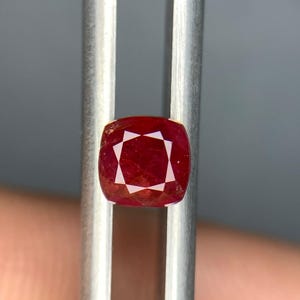 May include: A cushion-cut red gemstone held between the metal prongs of a pair of tweezers. The stone features a faceted surface that reflects light, displaying a deep crimson hue with visible internal clarity and a symmetrical geometric pattern.