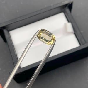 Puede incluir: Una piedra preciosa amarilla de corte cojín sostenida por pinzas de metal. La piedra presenta un tono amarillo pálido translúcido con facetas internas visibles que reflejan la luz. La gema se sitúa frente a una caja expositora negra con interior blanco.