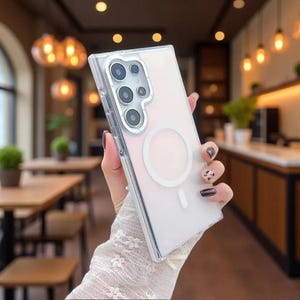 May include: A clear, transparent smartphone case with a white circular magnetic charging ring on the back. The case features a rectangular cutout for a multi-lens camera system, showing the silver-rimmed lenses of the phone inside the protective cover.