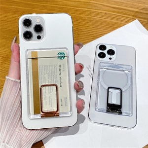 May include: Two white smartphones with triple-lens camera arrays. One phone features a clear card holder attachment containing a Starbucks card and a rose gold ring stand. The other phone has a clear magnetic ring attachment with a black folding stand.