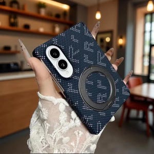 Può includere: Una custodia per smartphone blu scuro con un motivo a pixel bianchi ripetuto, un modulo fotocamera bianco che ospita due lenti circolari e un prominente anello magnetico nero circolare centrato sul retro della cover protettiva.