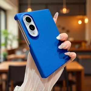 May include: A solid blue smartphone case with a matte finish, featuring a rectangular cutout for a dual-lens camera system. The case has a smooth, uniform texture and covers the back and sides of the device, leaving the camera lenses exposed.