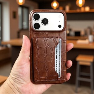 Puede incluir: Una funda de teléfono marrón con textura de piel de cocodrilo y un tarjetero integrado en la parte posterior. La funda cuenta con un recorte para las lentes de la cámara y sostiene una tarjeta de crédito con el texto AMERICAN EXPRESS visible.