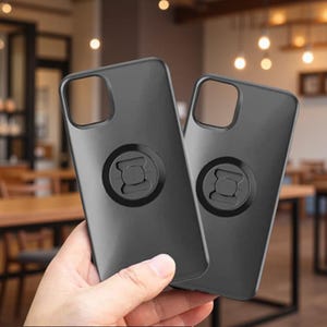 May include: Two matte black smartphone cases featuring a square camera cutout at the top left and a circular mounting mechanism centered on the back. The cases have a smooth, solid finish and are designed to fit a mobile device securely.