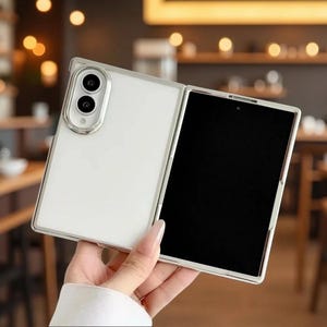 May include: A foldable smartphone with a white back panel and a silver-toned frame. The device features a prominent dual-camera module with a silver border on the rear. The front display is black and inactive, showing a sleek, modern design.