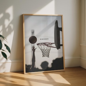 May include: A framed wall art print featuring a silhouette of a basketball being shot towards a hoop. The text NO EASY BUCKETS is printed in black near the centre. The image uses a high-contrast monochrome style with a light wooden frame.