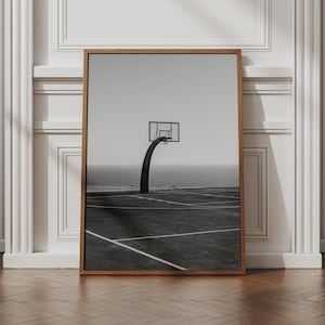 May include: A black and white framed wall art print featuring a basketball hoop standing on an outdoor court by the ocean. The scene shows a curved pole, a rectangular backboard, and white court lines painted on the ground surface.