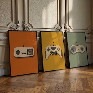 Op de afbeelding: Drie ingelijste rechthoekige wandkunstprints met illustraties van videogamecontrollers. De linkerprint heeft een oranje achtergrond, de middelste print is geel en de rechterprint is saliegroen, elk met een andere stijl controller.