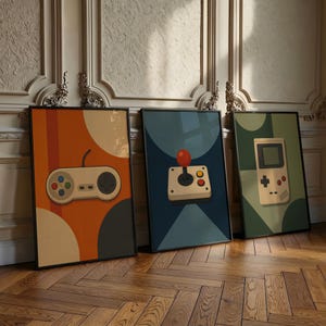 May include: Three framed rectangular wall art prints featuring retro gaming illustrations. The left print shows a beige game controller on an orange background. The center print displays a joystick on a dark blue background. The right print depicts a handheld game console on a green background.