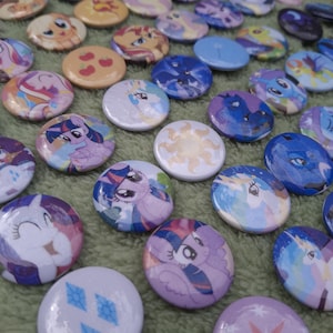 Könnte beinhalten: Eine Sammlung runder Ansteckbuttons mit verschiedenen bunten Cartoon-Pony-Figuren, Symbolen und Icons. Die Buttons zeigen vielfältige Illustrationen, darunter Pony-Gesichter, ein Sonnenemblem, Apfelmotive und Rautenformen in einer Vielzahl von Farben.