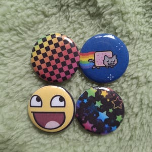 Op de afbeelding: Een set van vier ronde buttons. Eén heeft een regenboogkleurig geblokt patroon, een andere toont een gepixelde kat met een regenboogspoor op een blauwe achtergrond, één toont een geel lachend gezicht en de laatste heeft kleurrijke sterren op zwart.