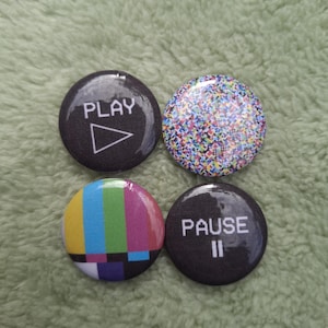 Op de afbeelding: Een set van vier ronde buttons. Eén is zwart met de witte tekst PLAY en een driehoekig icoon. Eén heeft een veelkleurig gepixeld patroon. Eén toont verticale kleurenbalken. Eén is zwart met de witte tekst PAUSE en twee verticale strepen.