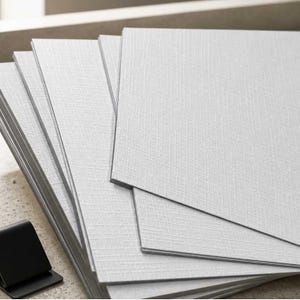 A stack of light linen textured rectangular panels or tiles, fanned out on a light stone surface with a small black stand in the foreground.