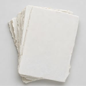 May include: A stack of rectangular sheets of off-white paper with uneven, deckled edges. The paper has a textured, fibrous surface and is arranged in a slightly fanned, overlapping pile, showcasing the raw, handmade quality of the edges.