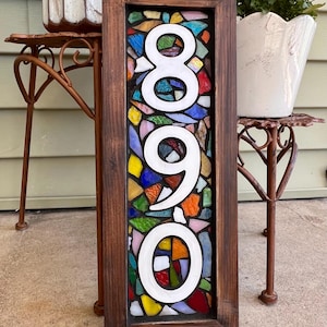 Pode incluir: Uma placa retangular de número de casa com os dígitos 8, 9 e 0 em branco, sobre um fundo de mosaico feito de peças de vidro colorido. A placa está inserida numa moldura de madeira castanho-escura com textura de grão de madeira visível.