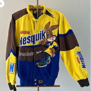 May include: A yellow, brown and blue varsity jacket featuring the Nesquik logo and the Nesquik bunny character. The front displays the text Nestle Nesquik chocolate flavour. The sleeves have a large N logo and the Nesquik brand name printed.