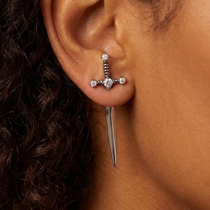 Peut inclure: Une boucle d'oreille en forme d'épée argentée dotée d'une lame pointue et d'une garde ornée de trois petites pierres rondes transparentes. La boucle d'oreille est conçue pour traverser le lobe, donnant l'apparence d'une épée transperçante.