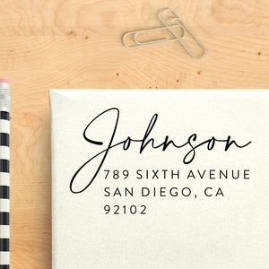 May include: A white envelope with the name "Johnson" in elegant black script, along with an address. A black and white striped pencil and two silver paper clips are also visible on a light wood surface.
