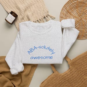 Peut inclure: Un sweat-shirt à col rond gris clair avec le texte bleu "ABA-solutely awesome" imprimé sur le devant. Le vêtement possède des manches longues avec des poignets côtelés et un col côtelé, présentant une texture de tissu chiné sur l'ensemble.