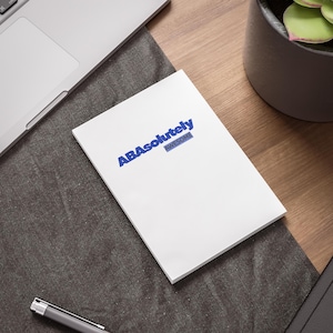 May include: A white rectangular notepad featuring the blue text "ABAbsolutely" with the word "AWESOME" printed in a smaller blue font directly underneath. The notepad is positioned on a dark gray textured mat next to a silver laptop and pen.