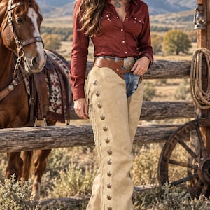 May include: A pair of beige suede chaps featuring long fringe along the outer legs and a vertical row of round silver-toned metal conchos. The chaps are worn over blue denim jeans, paired with a long-sleeved dark red button-down shirt and a brown hat.