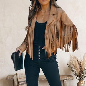 May include: A tan suede jacket featuring long fringe detailing along the chest, shoulders, and sleeves, paired with a black top and high-waisted black pants with a row of five visible buttons, accompanied by a black clutch held in one hand.