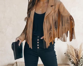 May include: A tan suede jacket featuring long fringe detailing along the chest, shoulders, and sleeves, paired with a black top and high-waisted black trousers with a row of five visible buttons, accompanied by a black clutch held in one hand.